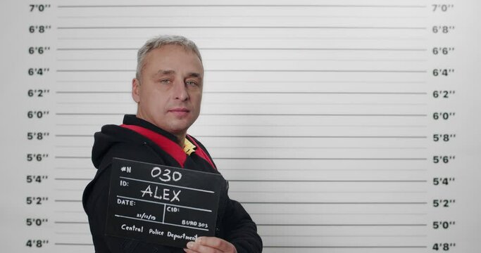 Side profile mugshot of male criminal holding sign while being photographed in police station. Arrested man in training suit turning head and looking to camera. Concept of crime.