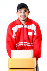 Happy Asian delivery man pushes stack of cardboard boxes on hand truck. Full length vertical portrait of courier guy in red uniform on isolated white background. E-commerce business concept.