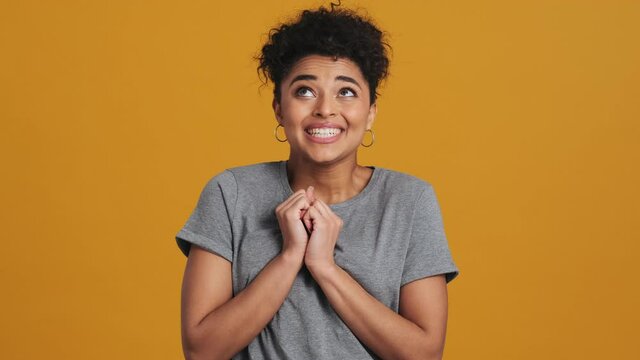A surprised emotional american woman wearing gray basic T-shirt is looking to the camera while rejoicing standing isolated over yellow wall in the studio