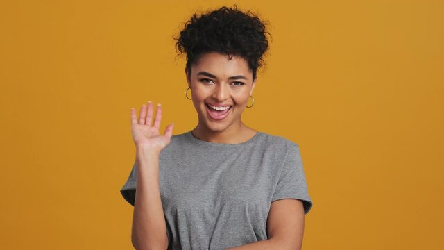 A positive american woman wearing gray basic T-shirt is doing hello gesture isolated over yellow wall in the studio
