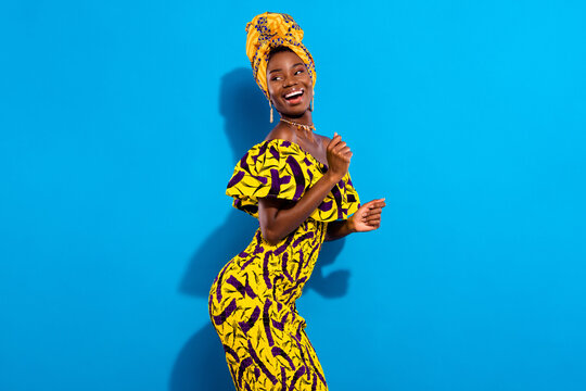 Photo of young smiling positive funky african girl dancing in yellow print dress look copyspace isolated on blue color background
