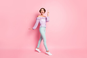 Full length photo portrait of woman walking showing v-sign isolated on pastel pink colored background