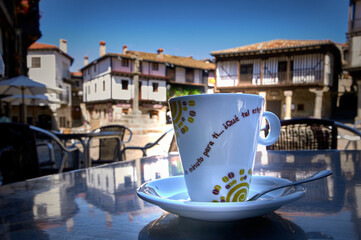 Fototapeta na wymiar Close-Up Coffee Cup On Metal Table On The Terrace Of A Bar With Townhouses Out Of Focus In The Background. On The Cup You Can Read...
