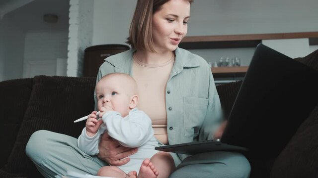 A positive mom writing something on a laptop and holding her little baby sucking pen while sitting on the couch