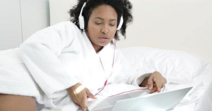 A cute African American woman in a white coat plugs headphones into a laptop and listens to an English lesson. Black woman study online during quarantine.