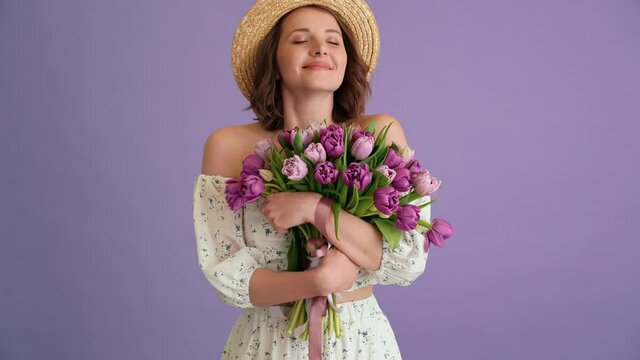 A positive woman with short hair in a dress and a hat enjoying the smell of a bouquet of tulips while holding it in her hands while standing in a purple studio