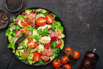 Fresh salad with salted salmon, avocado, egg, sesame seeds, olive oil, tomatoes and mixed herbs. Keto diet food. Black background, top view