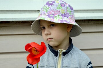 A pre-school boy sits on the threshold of the house, wearing a panama hat on his head and holding a...