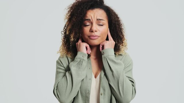 A displeased african american woman with afro curls is covering her ears with her fingers standing isolated over a white wall in the studio