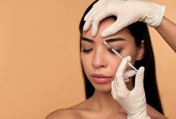 Young Asian woman with clean radiant skin gets botox injections for contour tightening, lip augmentation on a beige background. Spa care, facial skin care, beauty cosmetology.