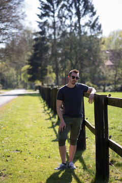 Gut aussehender Mann steht am Zaun und ist mit einer Hand dran abgelenkt. Ee ist Sommer und das Wetter ist sch&ouml;n, er hat Sonnenbrille auf und Sommersachen an.  Zaun ist lang. Im Hintergrund ist Wald