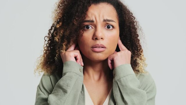 A shocked african american woman with afro curls is looking to the camera while covering her ears isolated over a white wall in the studio