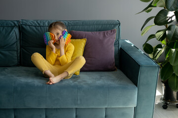 Obraz premium A little girl is playing with a modern popit toy. A fascinating sensory toy . Colorful and bright pop it toy. Simple dimple. The child is sitting on a sofa with yellow and purple pillows.