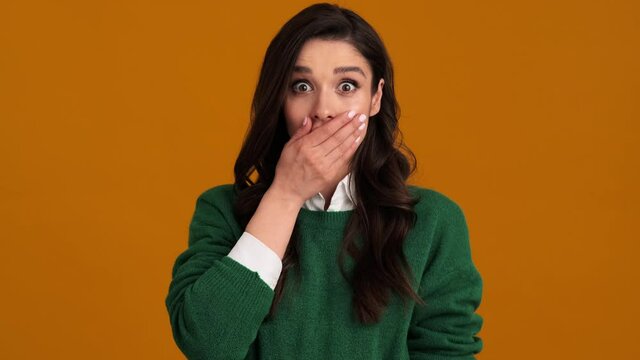A shocked woman in a green sweater and shirt saying wow and covering her mouth while standing in an orange studio