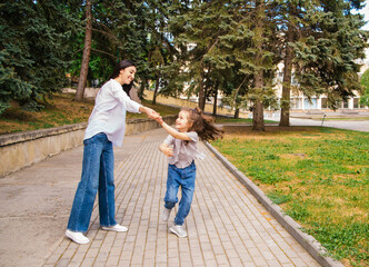 Naklejka na ściany i meble A young mother and her little daughter are dancing in the park. A girl and a woman are circling on the sidewalk.
