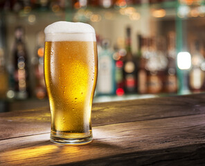Cooled glass of pale beer with condensation drops on glass surface on the wooden table. Blurred bar at the background.