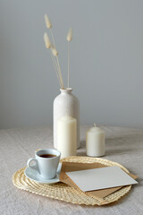 White paper card mockup, coffee cup, candles, vase of dried flowers on table. Romantic breakfast concept. Boho style.
