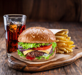 Delicious burger with cola and potato fries on a wooden table with a dark brown background behind. Fast food concept.