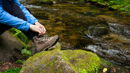 Female sitting on a mossy green rock by a stream mountain river fixing lace with hands on a brown...