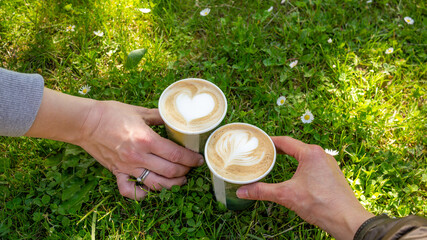 Hands holding a take away coffee cappuccino cups with milk foam heart  decoration over green and daisy background. Valentine Day. Coffee break in the park, little moments Cheering friends, lovers