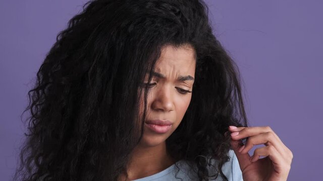 A close-up shot of disgruntled african woman looking at split ends while standing in purple studio