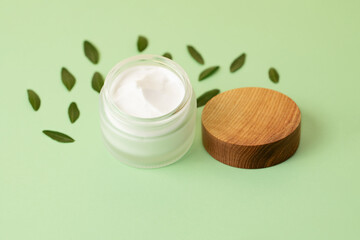 Open skin care packaging with white cream and wooden cover near it.Trendy mint background with copy space,front view.Concept of the zero waste,organic cosmetic.Fresh green leafs around it.