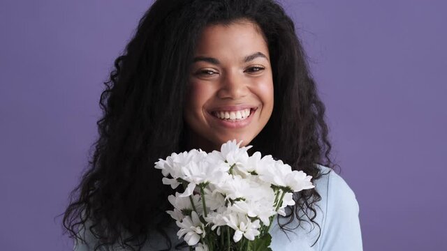 A smiling african woman in a t-shirt enjoying the smell of the flowers while standing in a purple studio
