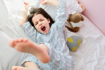 Top view of a little girl in white-blue striped pajamas, raising her legs directly to the camera and stretching out in a white bed, yawning, waking up early in the morning in a sunny bedroom.