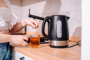 A black kettle is on the kitchen table. A woman's hand holds a teapot and makes tea. Tea bag in a...