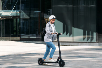 Young businesswoman with a safety helmet on her head driving electrical scooter near an office building