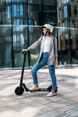 Side view young woman standing on electrical scooter near an office building. Female in helmet...