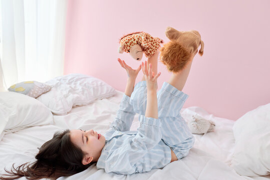 Indoor image of a playful little girl in pajama playing with toys on the bed with white bedding, on pink background. Child playing in the morning in her sunny bedroom.