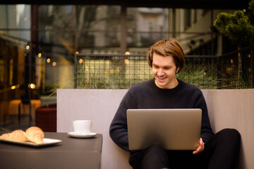 Man drinking cup of coffee in a restaurant on the terrace. Freelancer outdoor with a laptop, has breakfast with croissants. Businessman looks and dreams. Man with laptop . headphones.