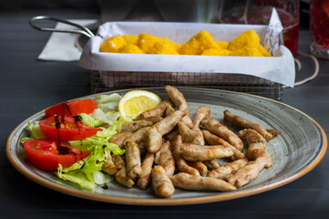 Lemon slice, lettuce, tomatoes and baked chicken sticks in a plate, breaded cheese in the background