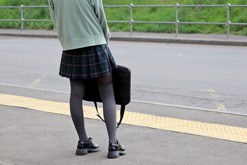 School girl in stockings and plaid skirt standing with laptop bag on bus stop