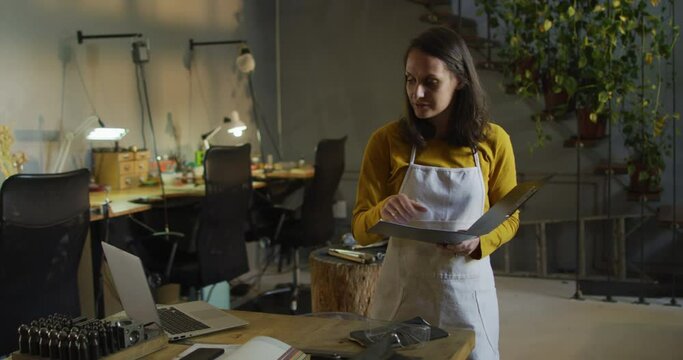 Caucasian female jeweller in workshop wearing apron, making notes, using laptop