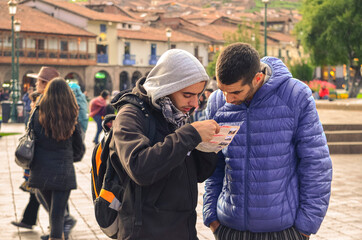 Fototapeta premium Two young male tourists holding and checking a map in travel destination city. Main city square (barrack square) of Cusco in Peru. Friends walking tour in cold morning
