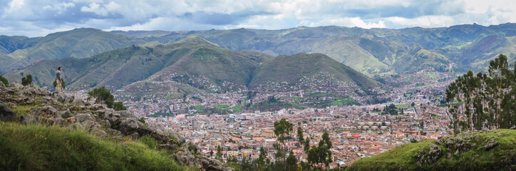 Fototapeta premium Panoramic view of a young man on top of a hill contemplating Cusco city landscape from the Sacsayhuaman inca archaeological site valley and ancient fortress. Peru, South America