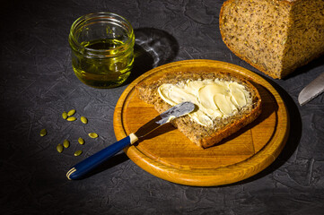 Slice of rustic natural yeast-free bread with flax, poppy seeds, sesame seeds, millet, pumpkin and sunflower seeds, spread with butter, on a wooden board, with olive oil in a glass jar