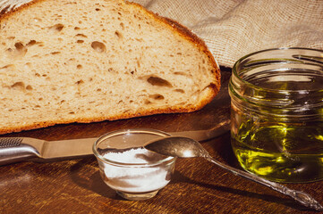 Still life - yeast-free buckwheat bread with olive oil and coarse salt in glass jars, a knife, and a linen napkin on a wooden board, wooden background, hard light, photo in a low key.