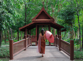 back side of Asian woman in Thai traditional dress holding red umbrella walking on bridge into...