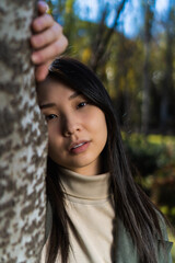 young multiracial asian american woman, posing behind a white tree, in a city park.