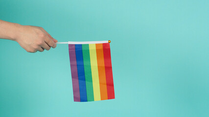 Hand is holding a rainbow flag on mint green or tiffany blue background.
