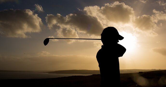 Composition of silhouette of golf player over landscape and clouds on blue sky with copy space