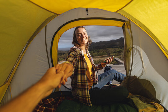 Smiling caucasian woman camping, sitting outside on mountainside deck holding hand of friend in tent
