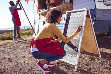 Diverse couple opening and preparing food truck by seaside on sunny day, woman writing on menu board