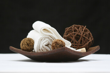 kit de decoración para sesión de spá aislado apoyado sobre mesa blanca y con fondo negro. Toallas, jabón, bolas de mimbre de ratán decorativas sobre plato de madera rústica.  