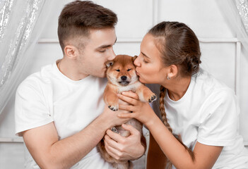 A happy couple kisses their cute red puppy shiba inu on the couch at home in the living room. Happy cozy moments of family life.