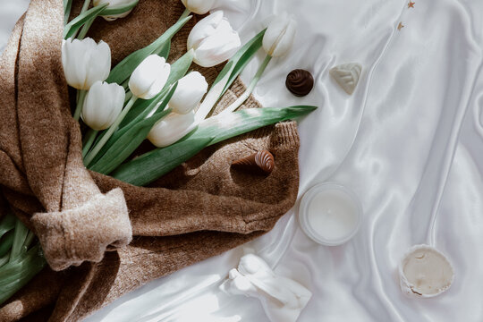 Bunch of white tulips wrapped in a woolly sweater on a silk tablecloth with candles