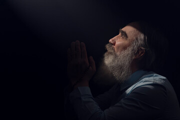 A senior man standing in dark smoky background light ray from above. Prayer gesture - obrazy, fototapety, plakaty